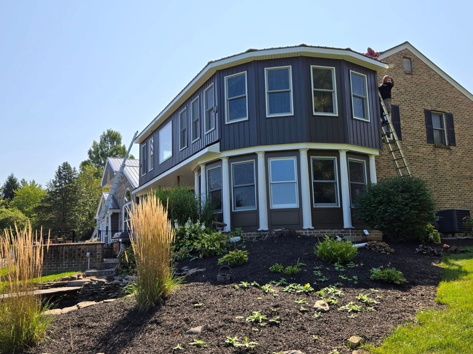 Two-story house with dark gray siding, numerous windows, and landscaping; person on a ladder.