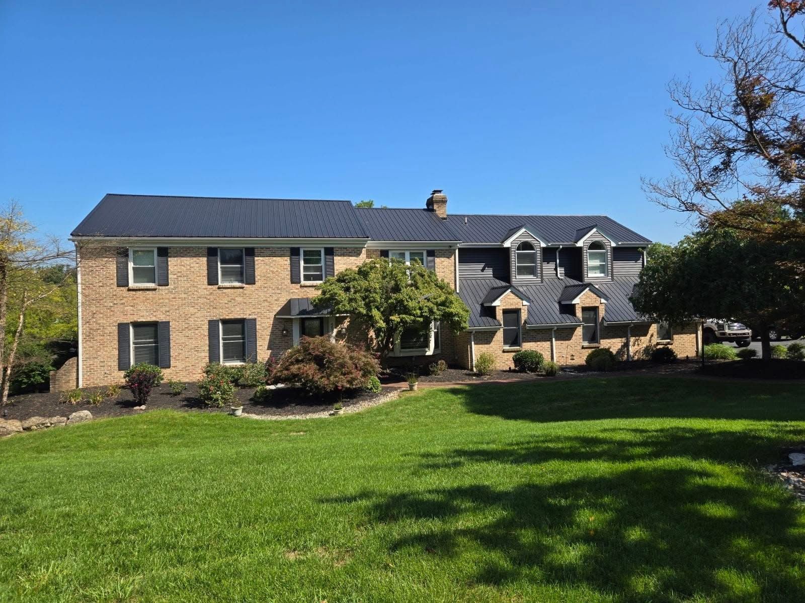 Two-story brick house with a dark roof and manicured lawn under a clear, blue sky.