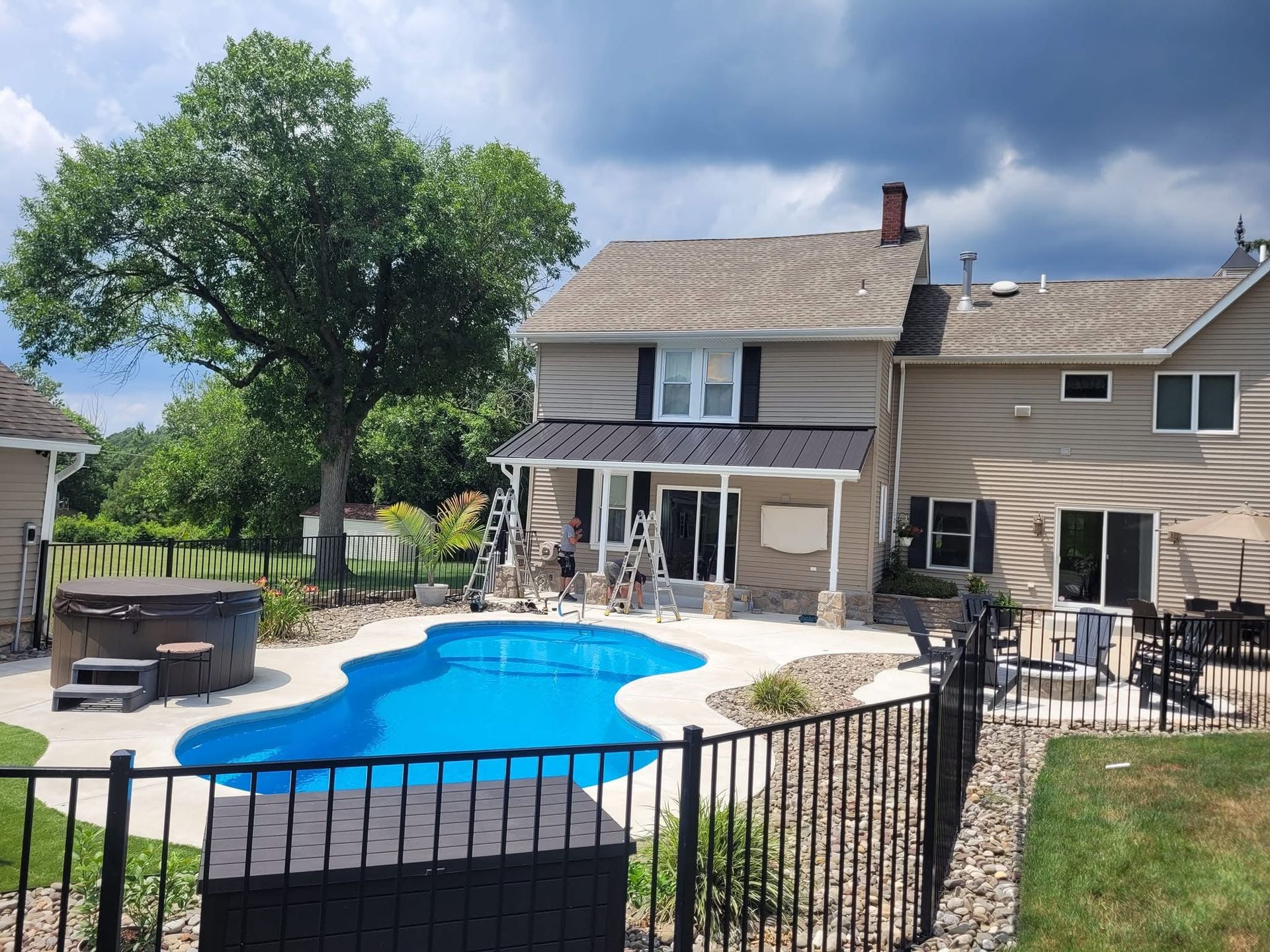 Backyard with pool, hot tub, and house. Blue pool, black fence, and tan house under a cloudy sky.