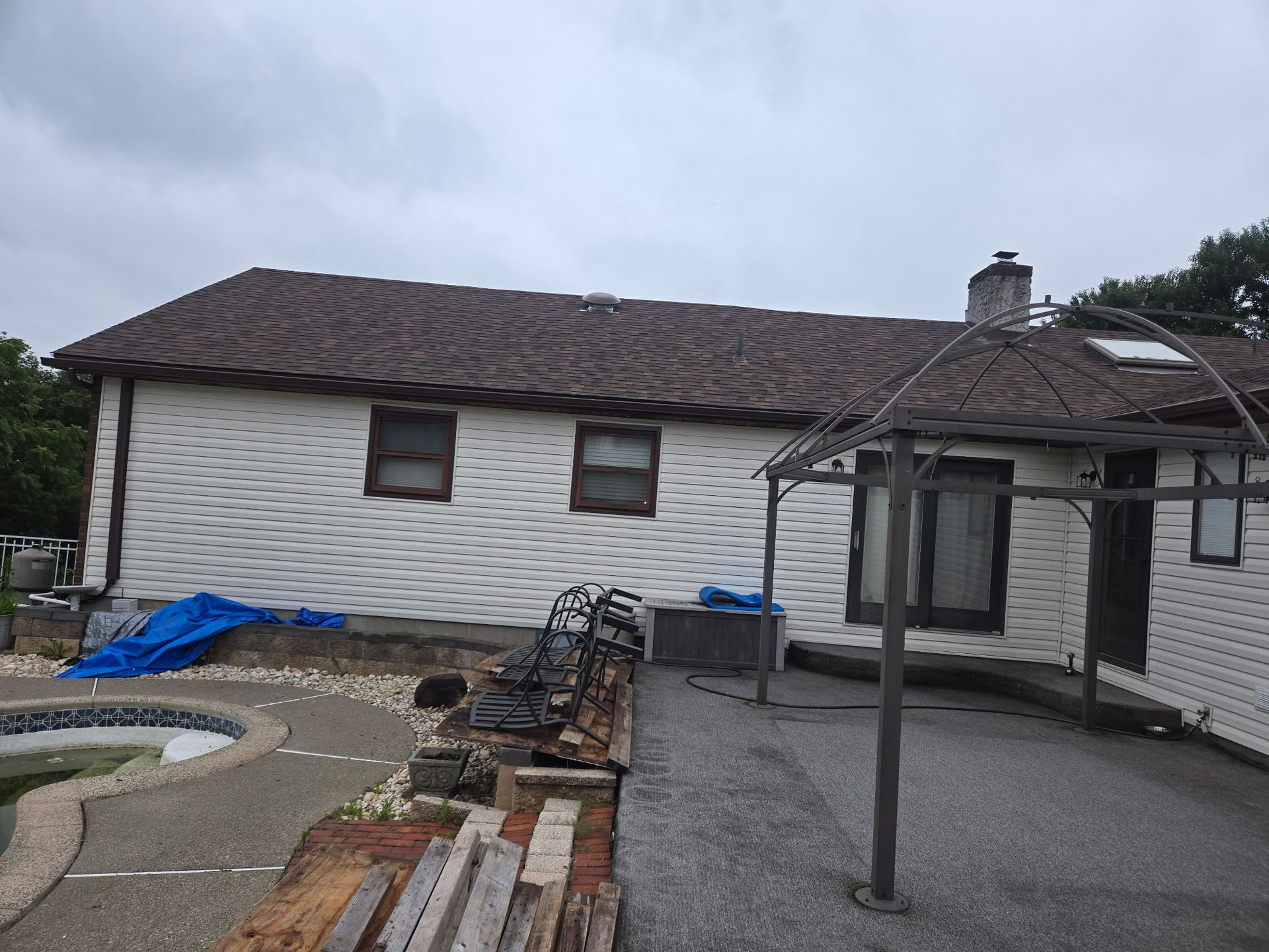Back of a white-sided house with a brown roof and a backyard patio and pool on an overcast day.