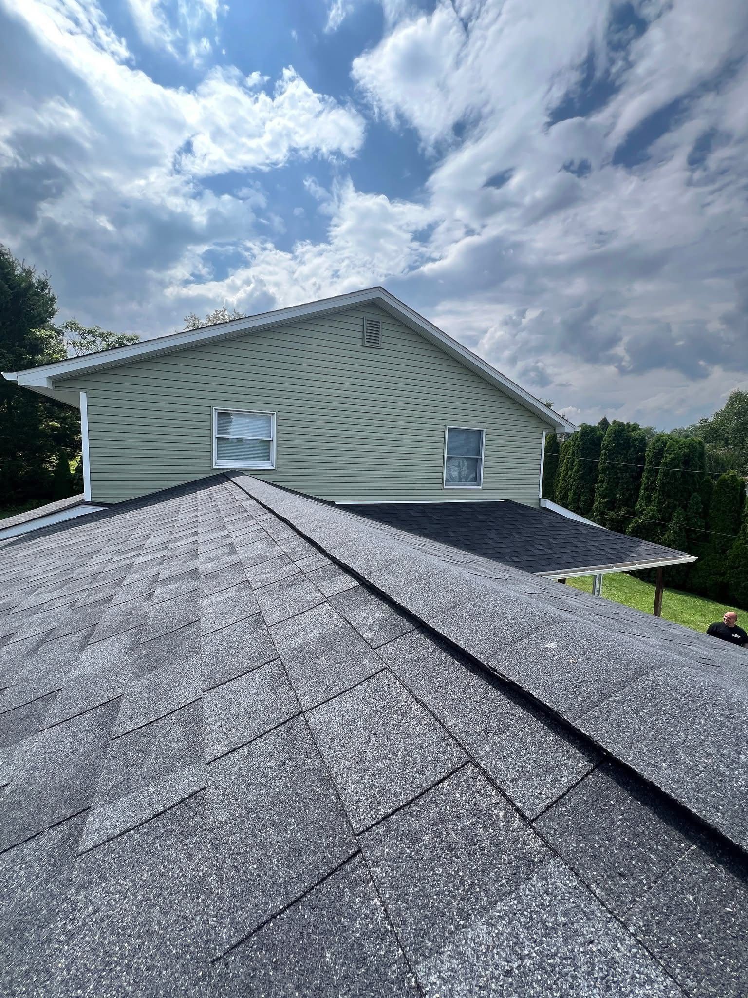 A house with dark asphalt shingles, light green siding, and two windows. Cloudy sky overhead.