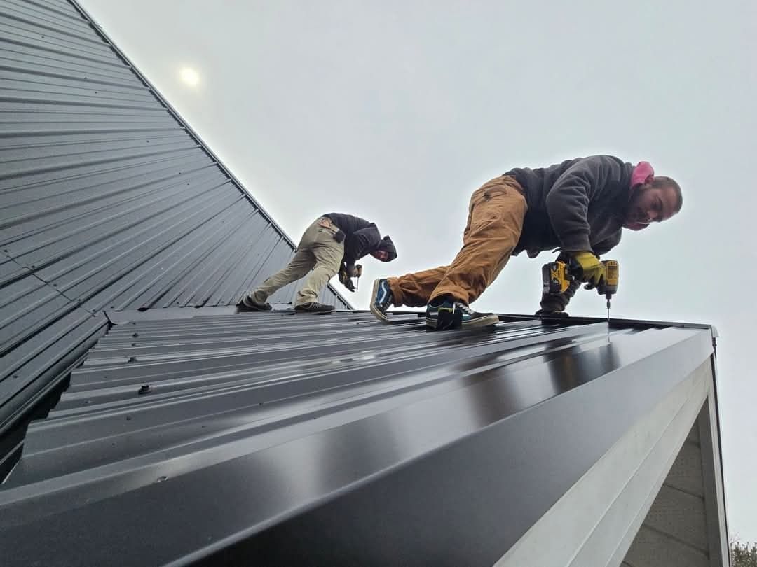 Two workers installing black metal roofing on a cloudy day.