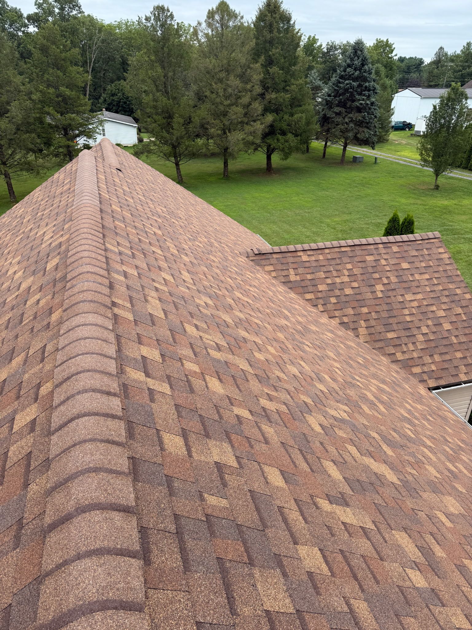 Brown shingled roof with a peak. Green trees and grass in the background. Overcast sky.
