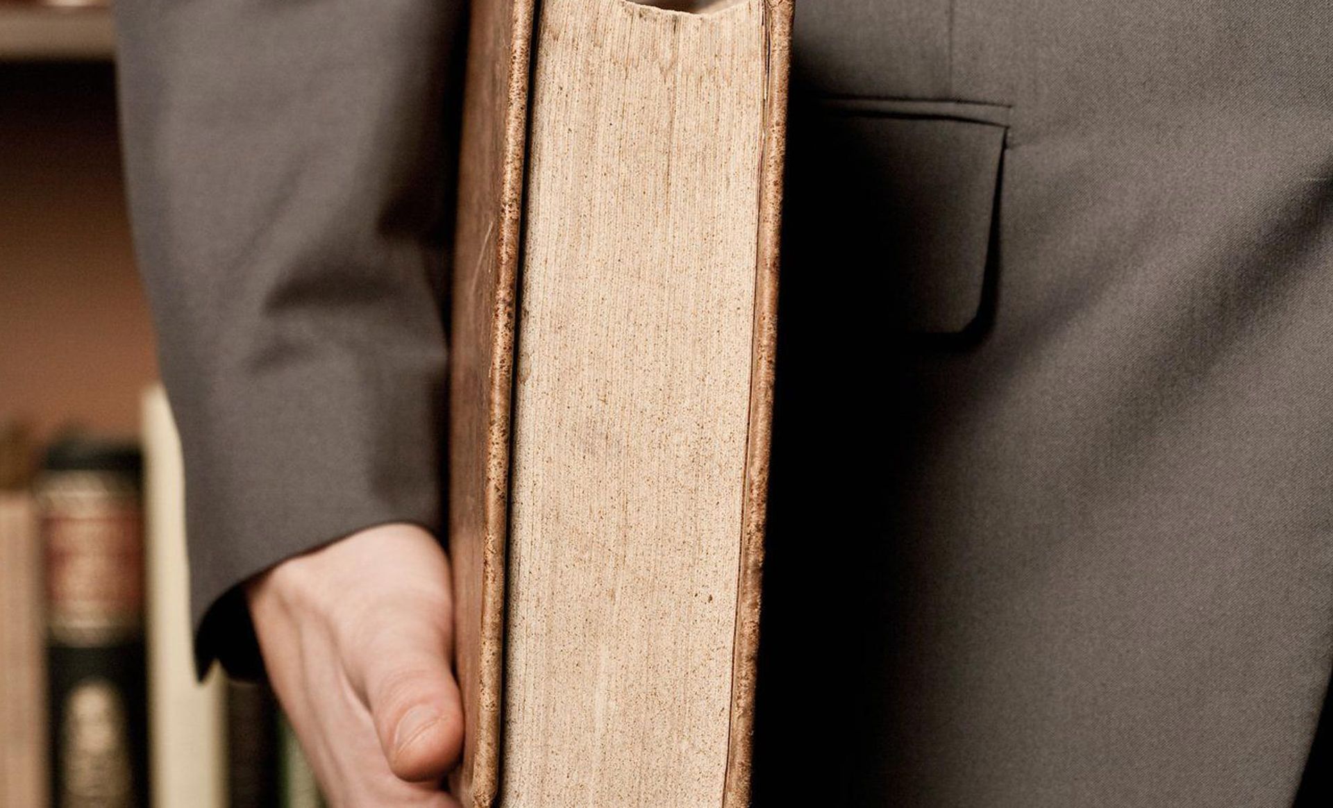 A man in a suit is holding a large book in front of a bookshelf.