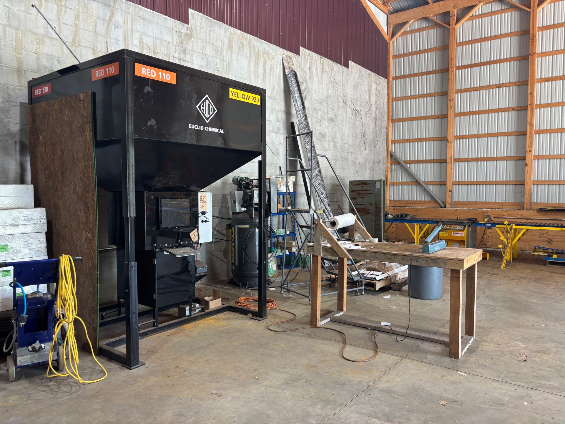 Large black industrial machine in a workshop beside a metal worktable and laddered storage area