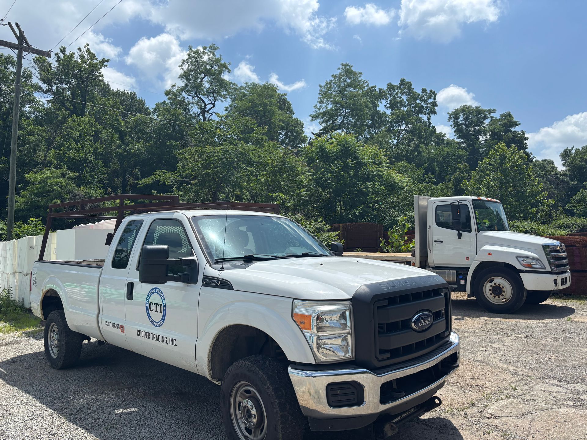 Two white utility trucks parked on gravel under a sunny, tree-lined sky.
