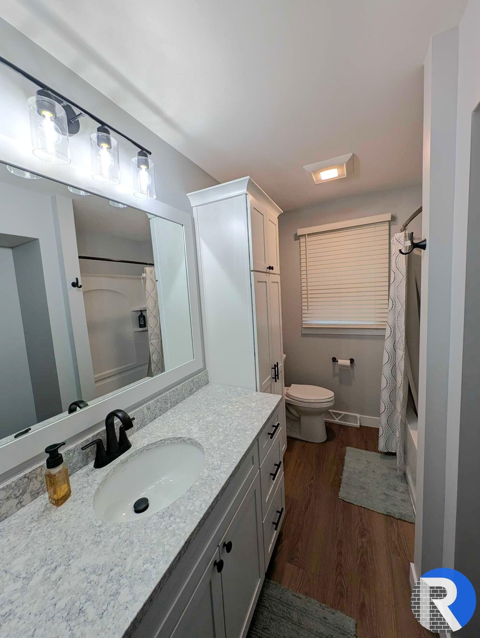 Bright white bathroom with double-sink vanity, large mirror, toilet, window blind, and gray rug.