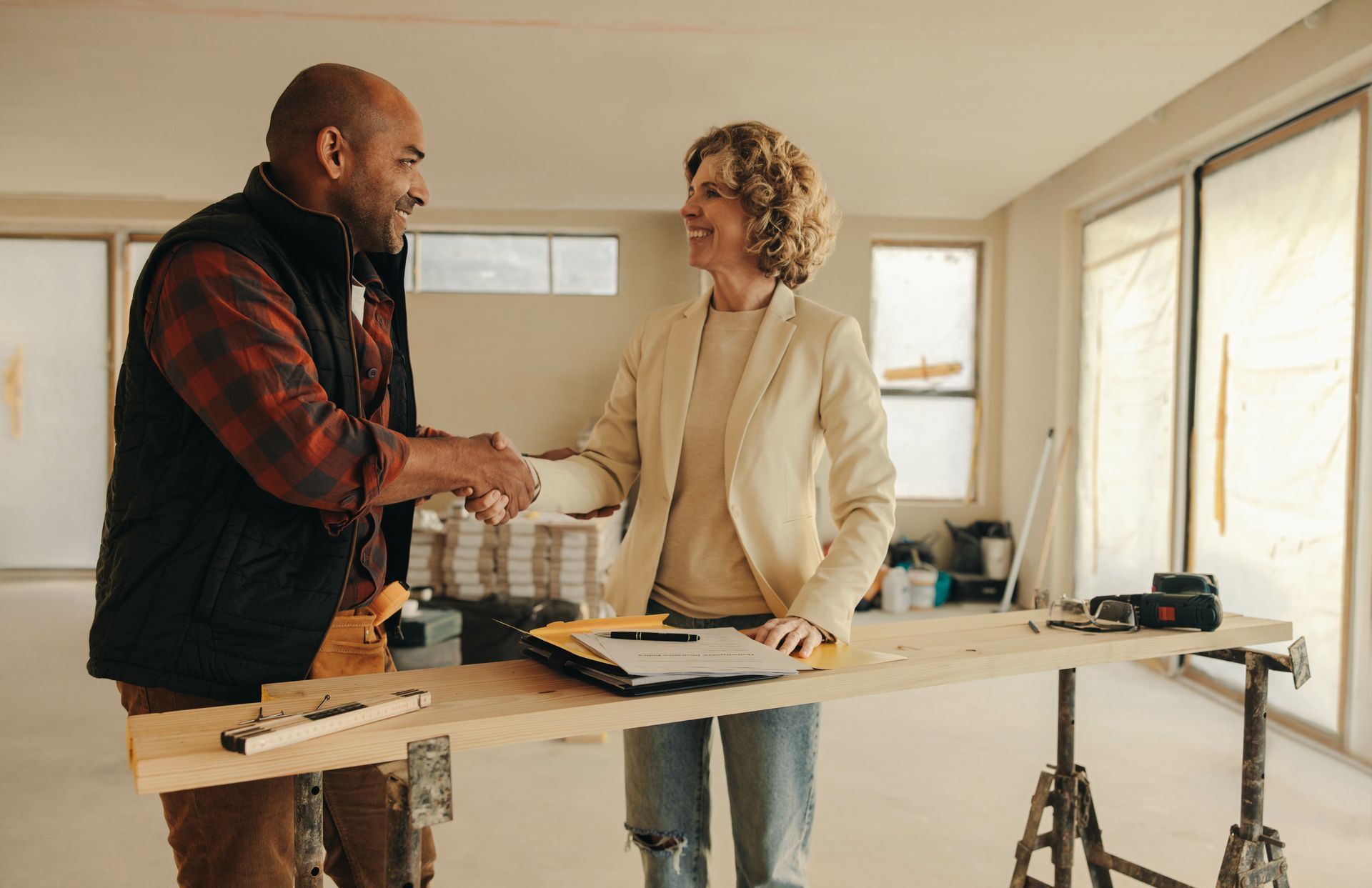 Two people shake hands in a bright, unfinished room with tools on a workbench.