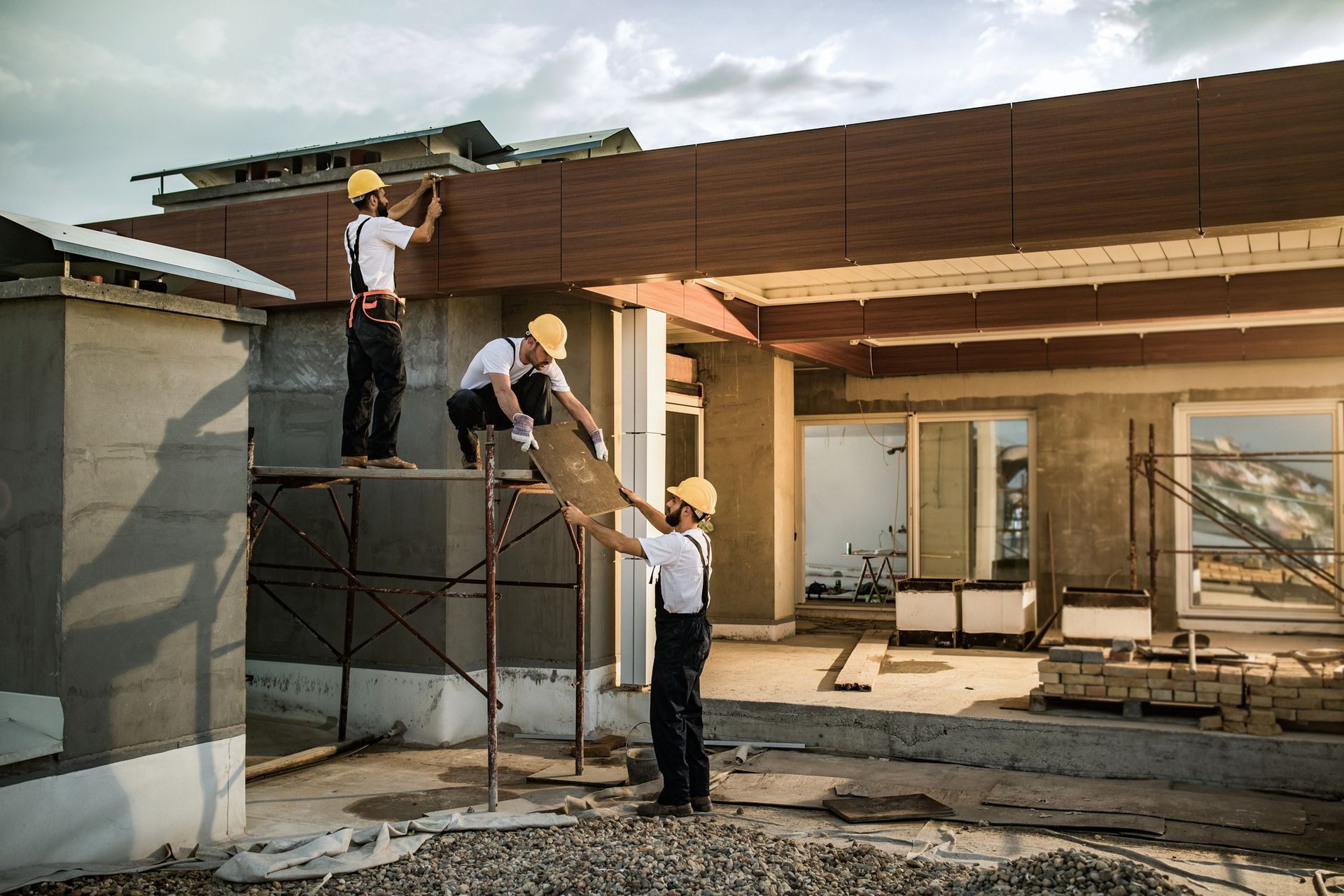 Workers building a modern house frame, one on scaffolding and two passing materials at the entrance.