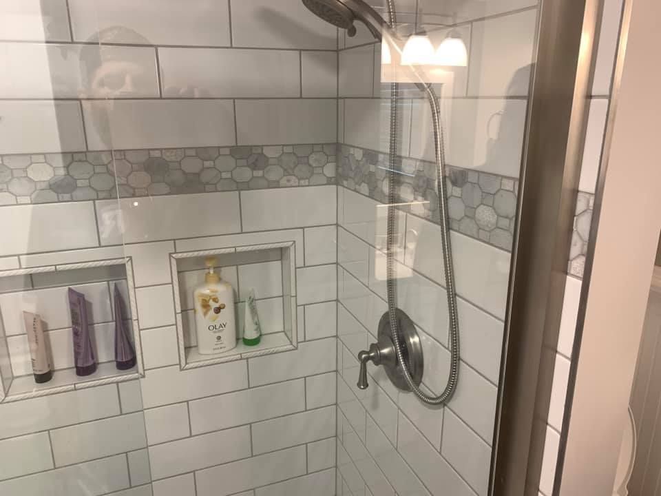 White-tiled shower with glass door, built-in shelves, and a silver handheld showerhead.