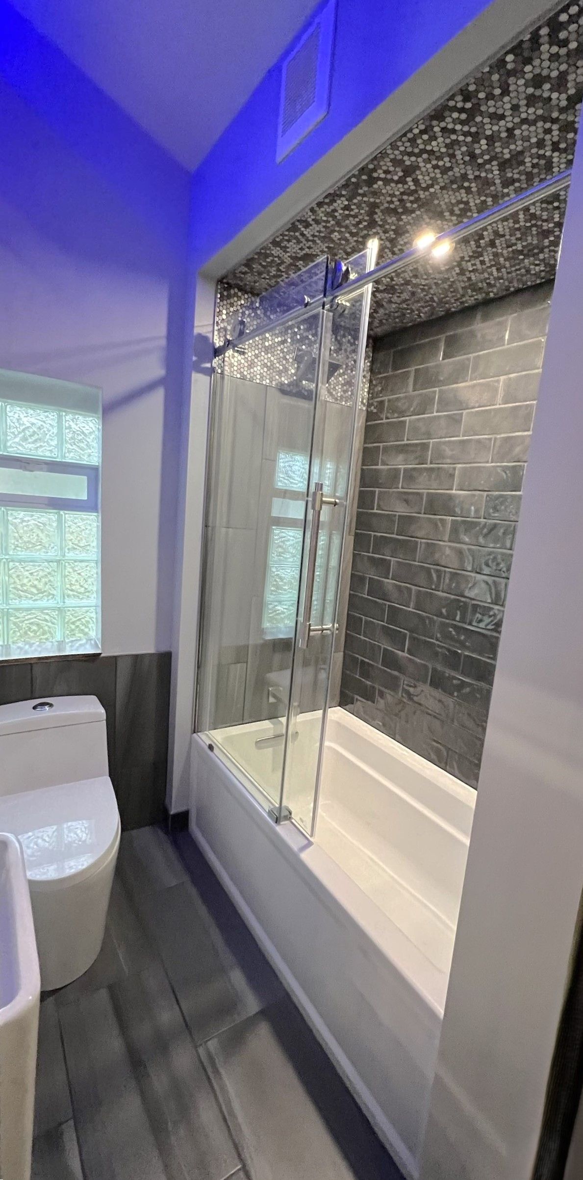 Modern bathroom with glass shower enclosure, dark tiled wall, and blue ceiling lighting