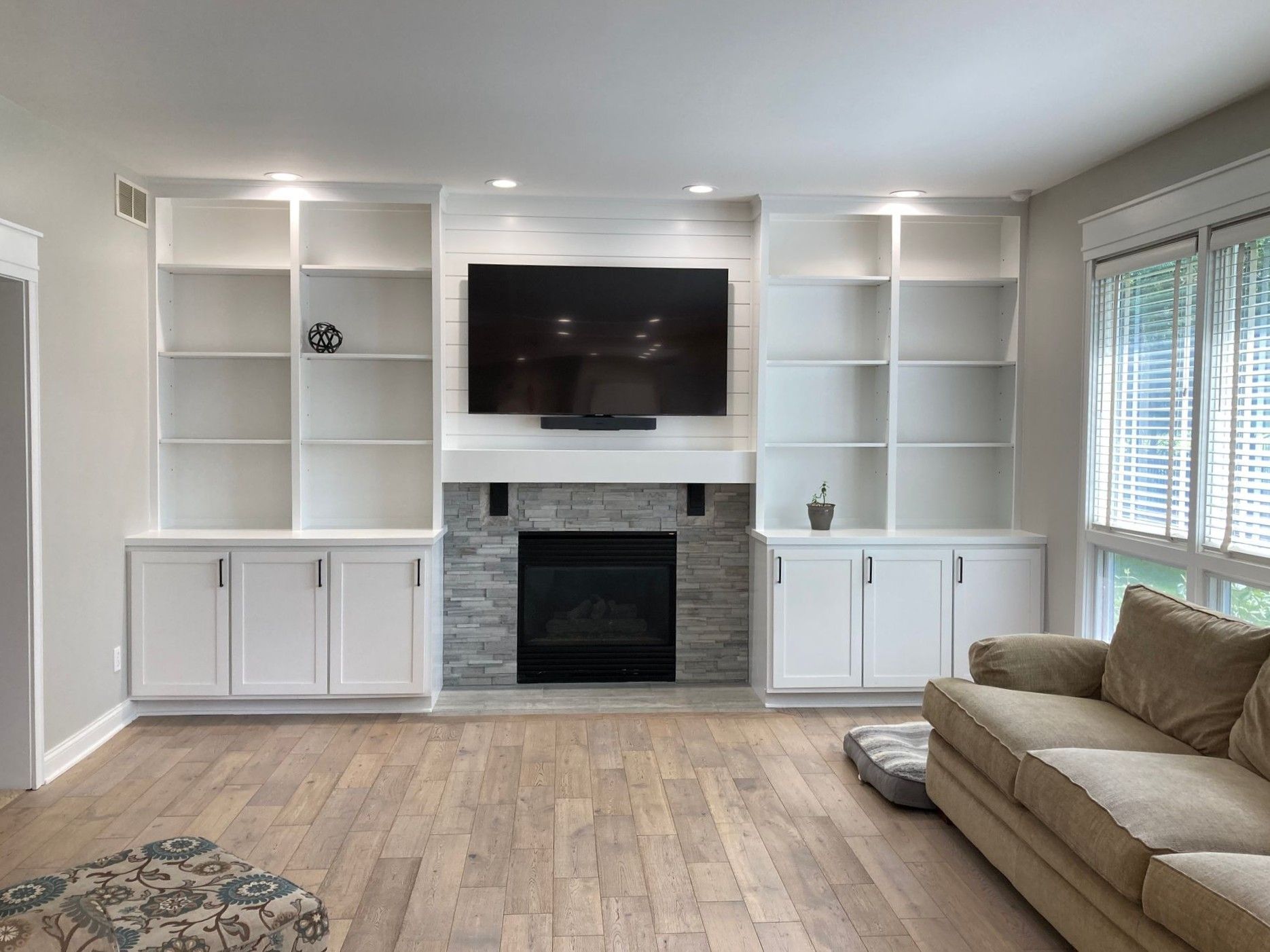 Bright living room with built-in white cabinets, stone fireplace, wall-mounted TV, and beige sofa