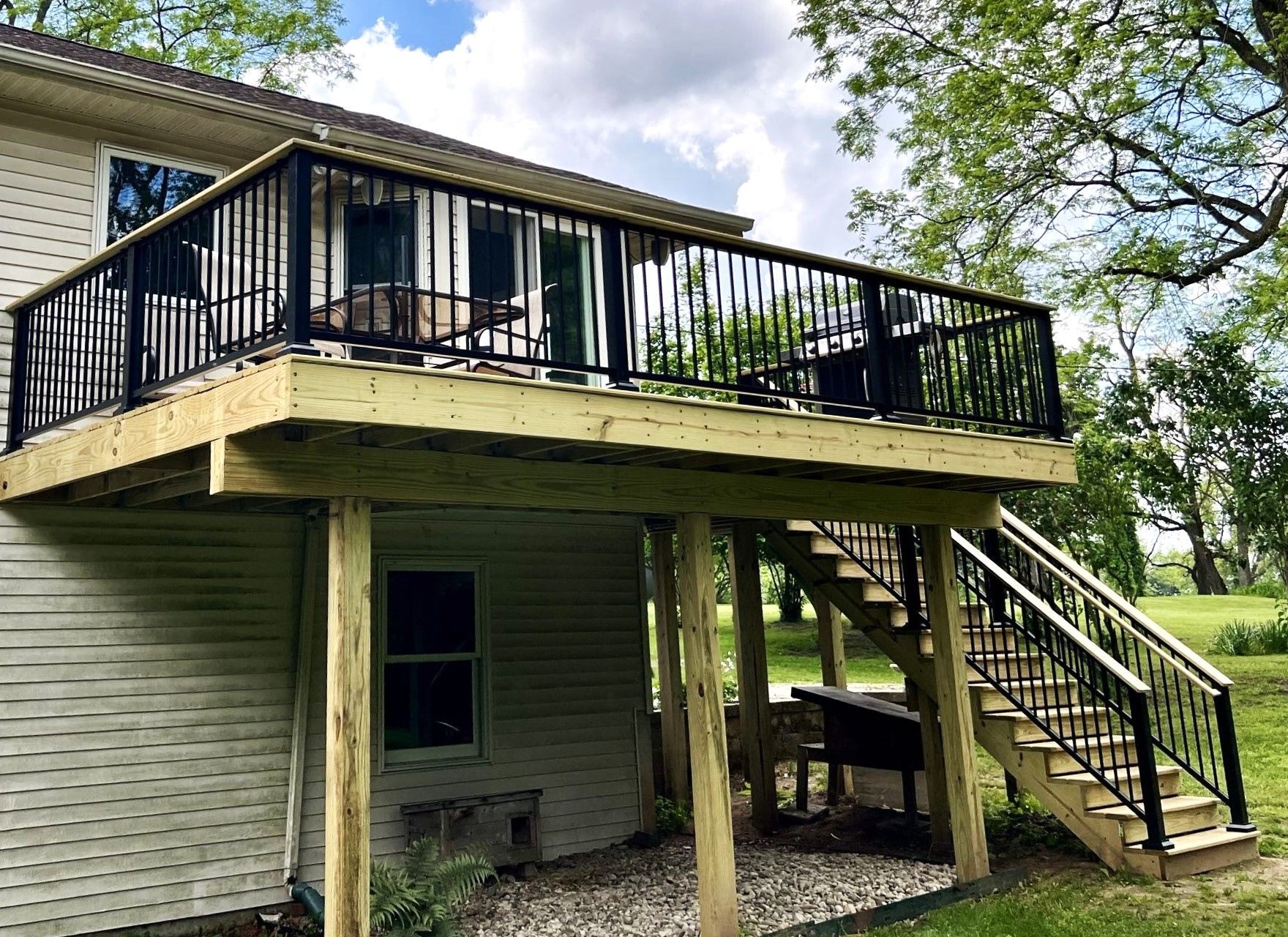 Raised wooden deck on a house with black railings and stairs, viewed from below in a grassy yard