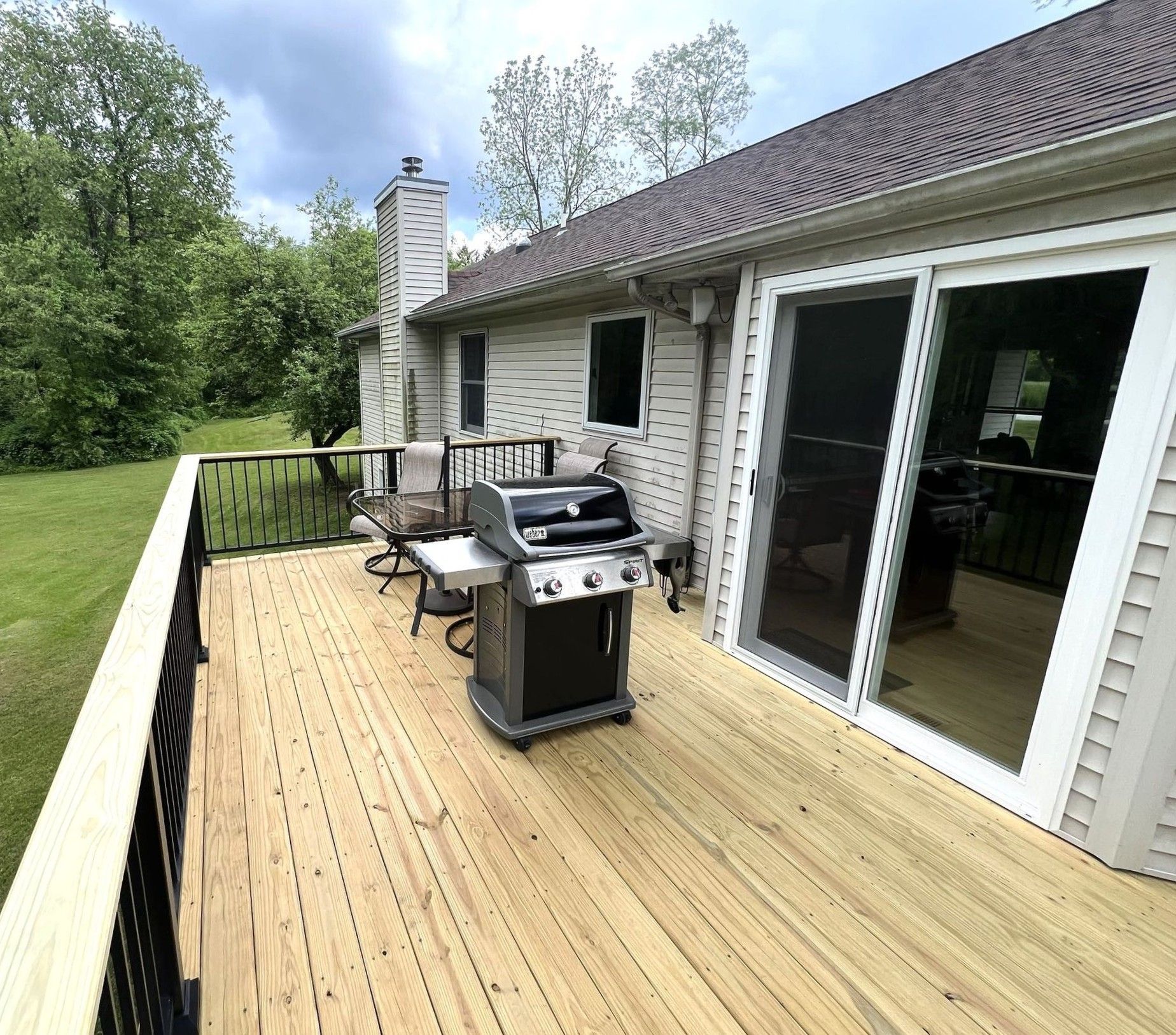 Back deck with grill, patio furniture, and white sliding doors beside a house, bordered by trees
