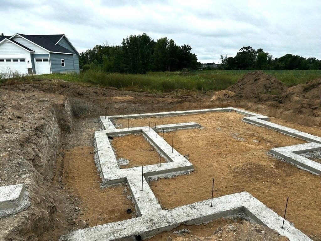 Concrete foundation footings in a dug-out lot beside a white house and grassy field