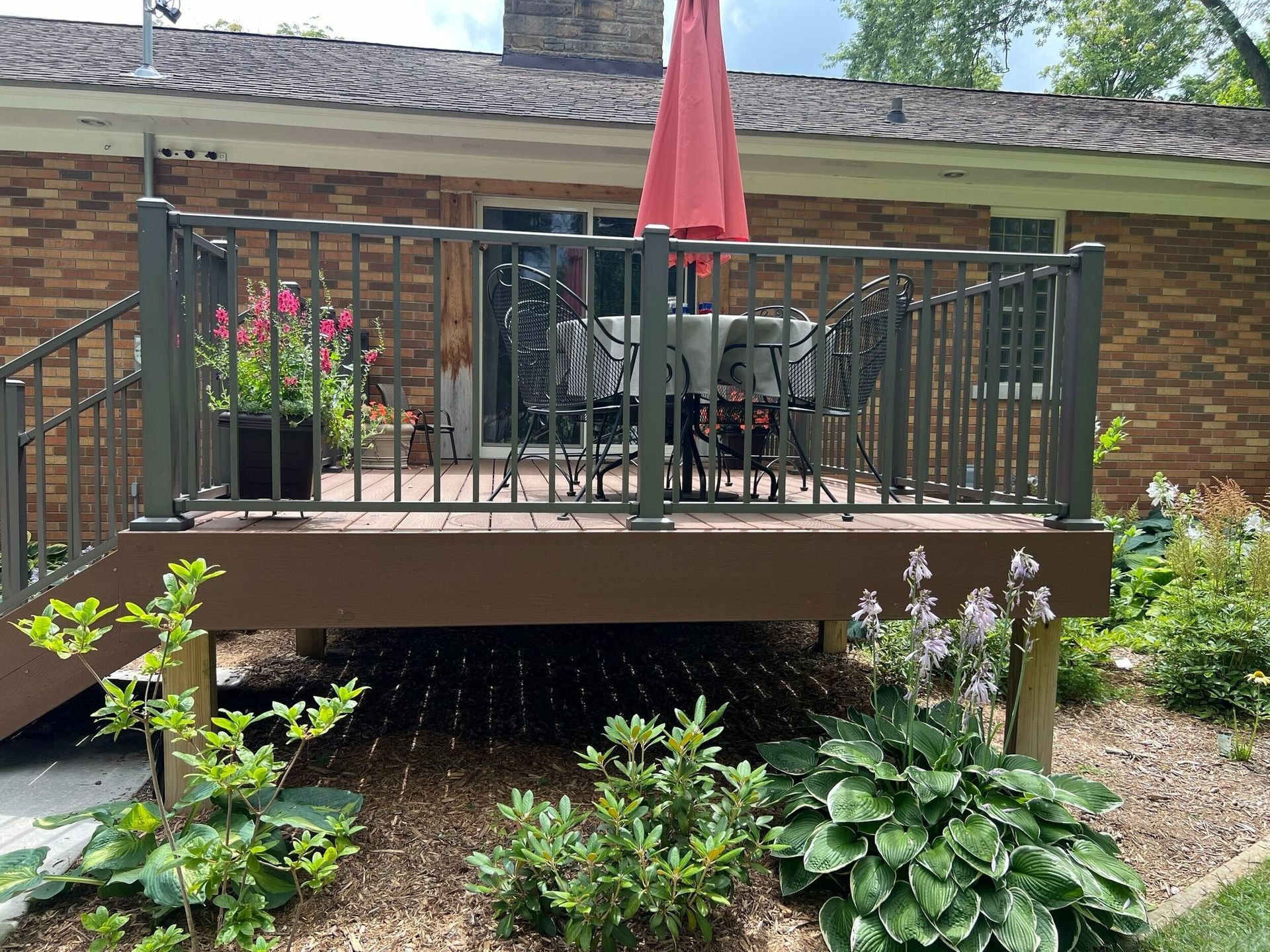Backyard patio with black railing, red umbrella, and brick house, surrounded by garden plants