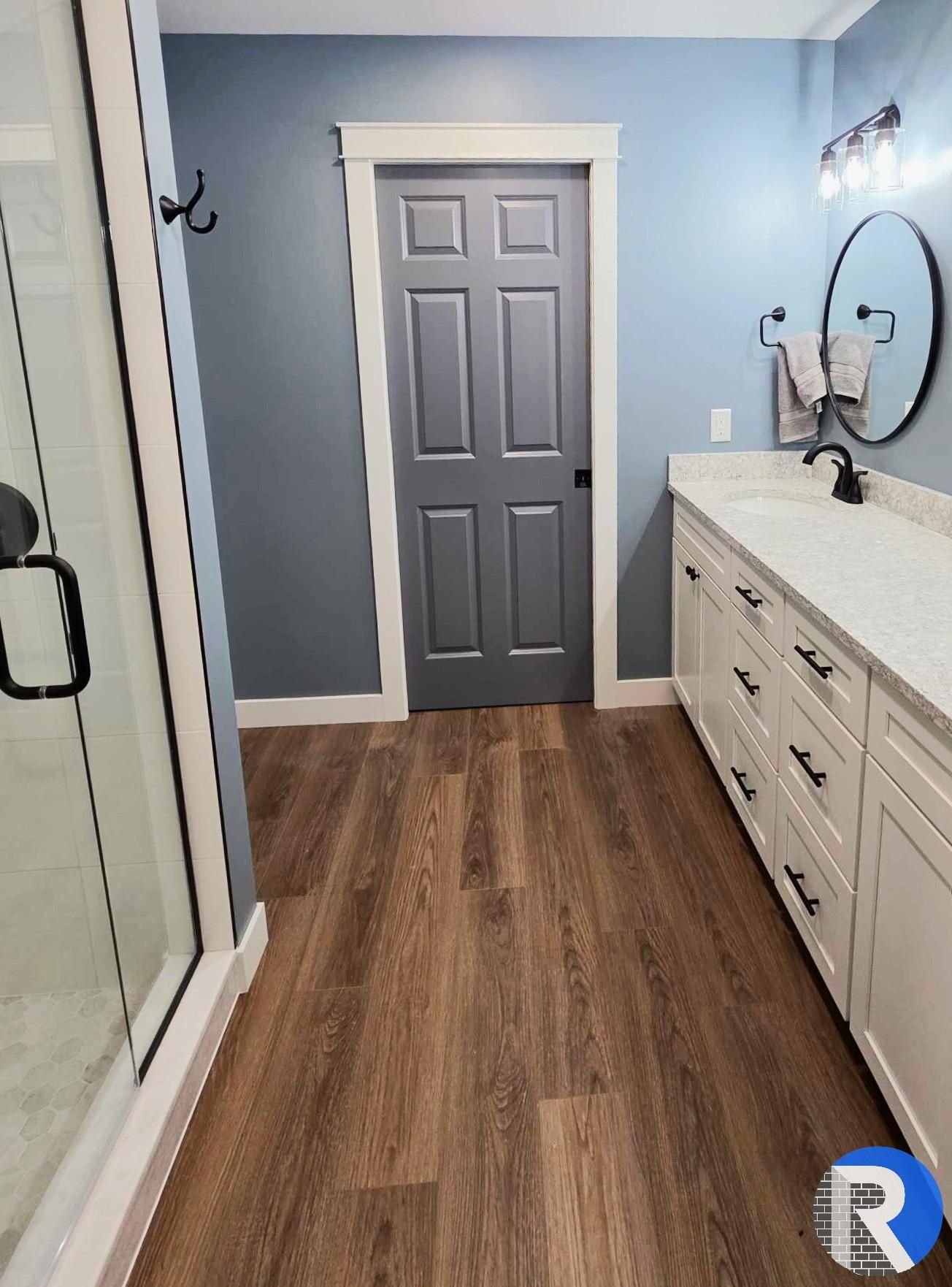 Bathroom with blue walls, gray door, wood floor, glass shower left, and white vanity with round mirror right
