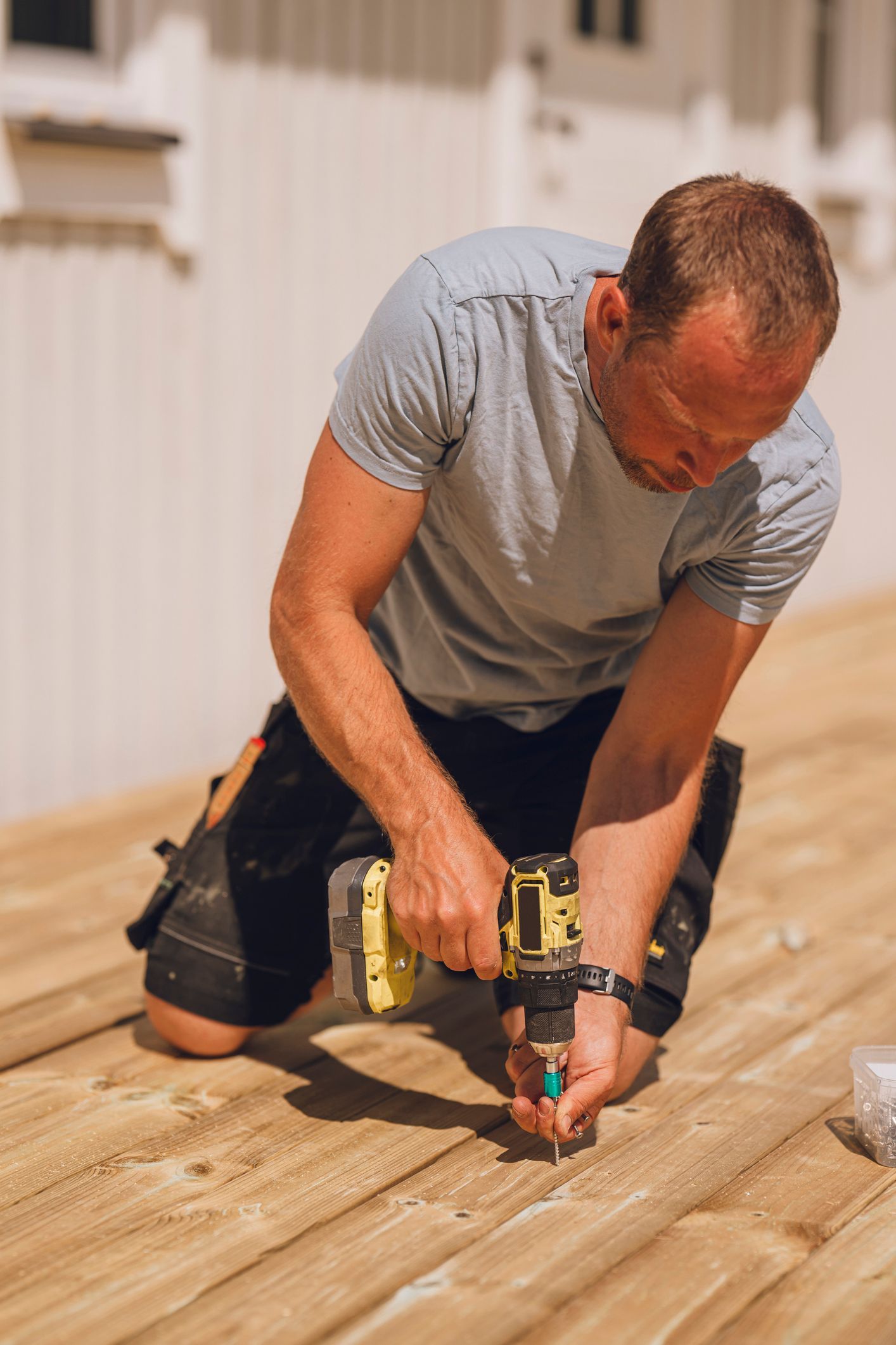 Man kneeling on a wooden floor, using a cordless drill to install flooring planks