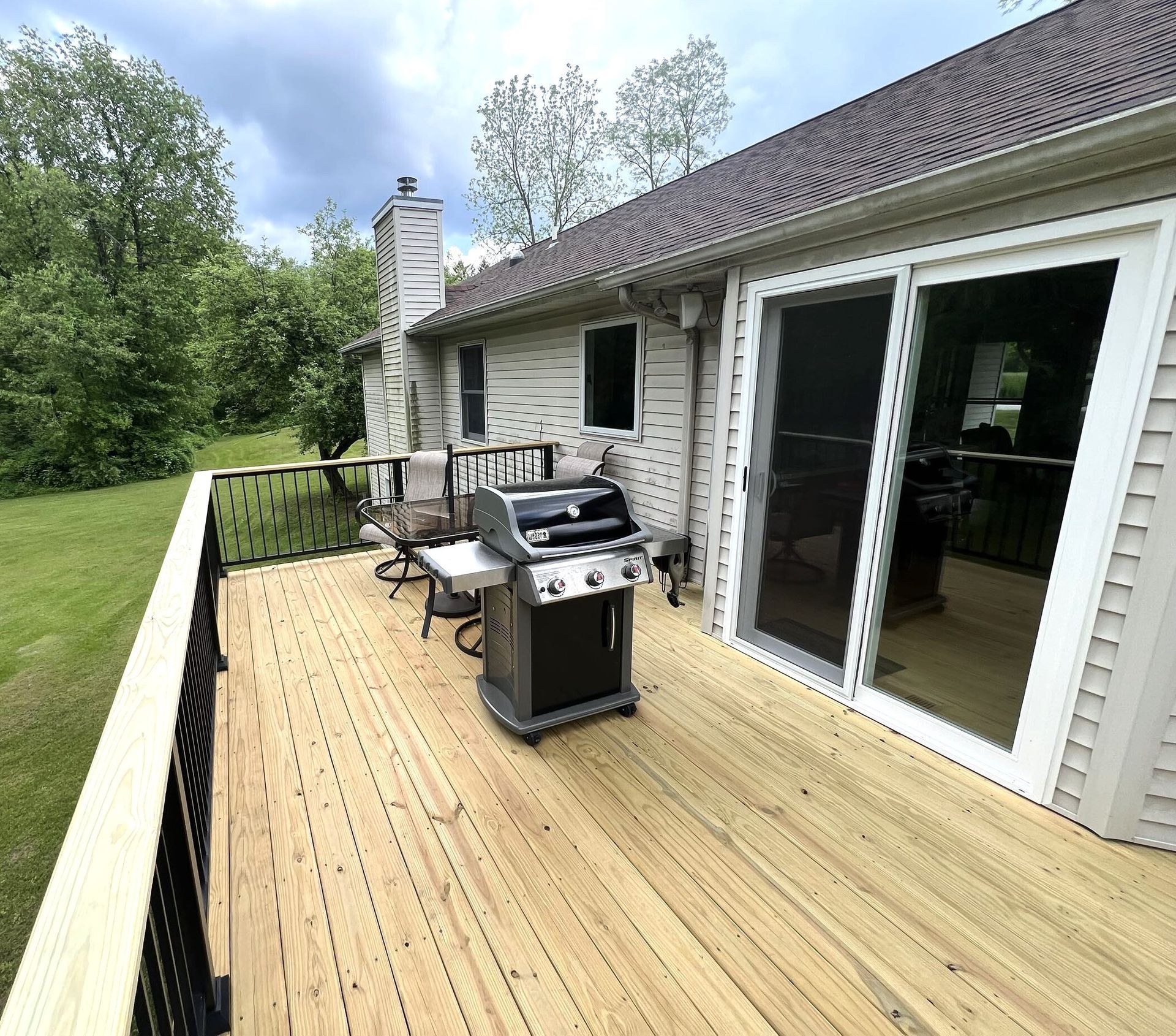 Wooden backyard deck beside a white house, with grill, chairs, and black railing overlooking trees.