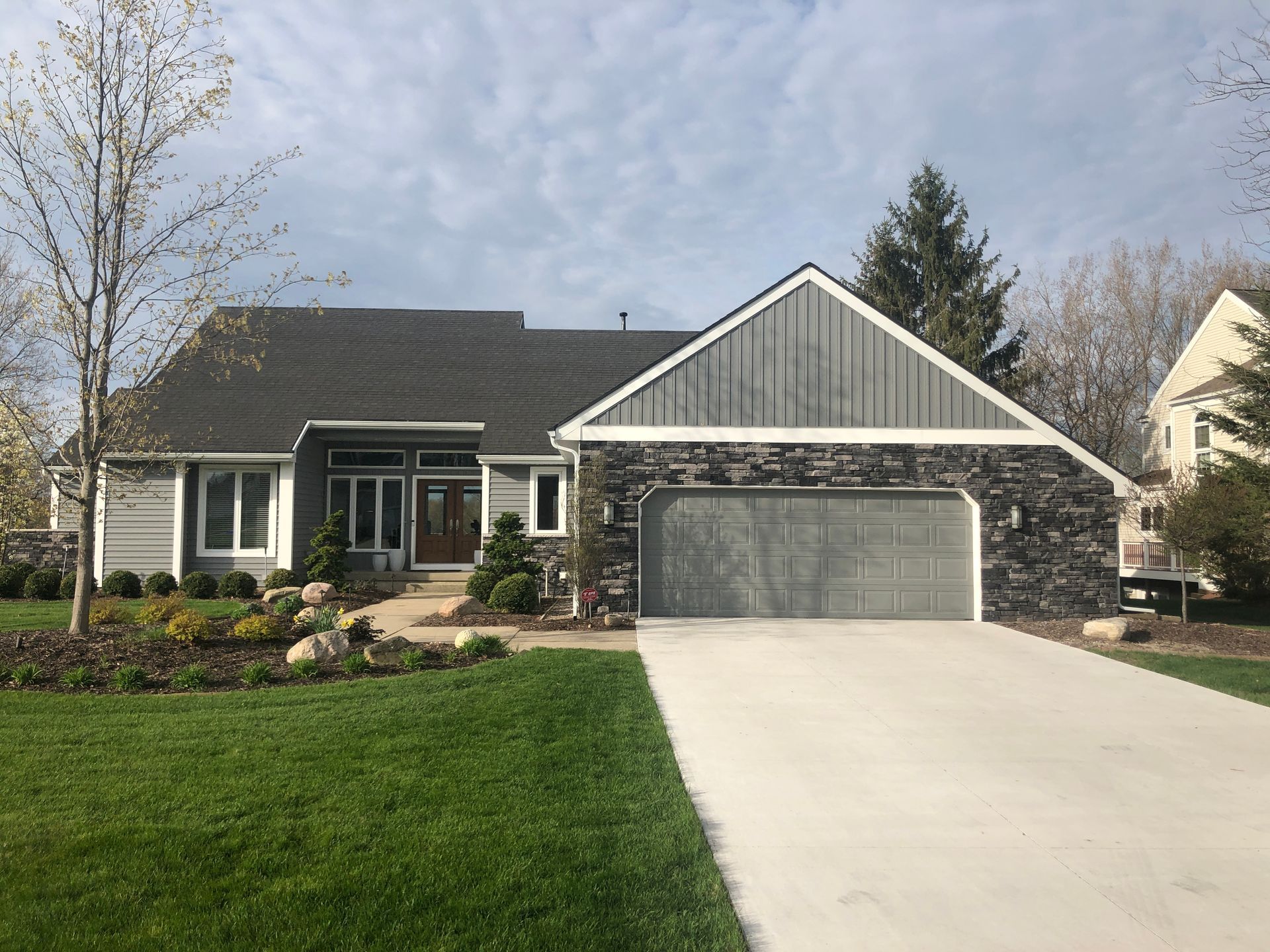 Suburban house with gray stone exterior, two-car garage, and a manicured lawn under a partly cloudy sky