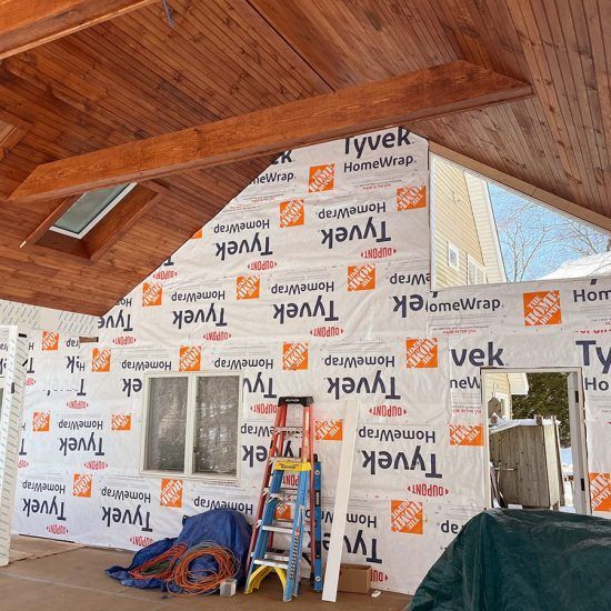 Interior of a wood-ceilinged cabin under renovation, with Tyvek wall wrap and ladders against the wall.