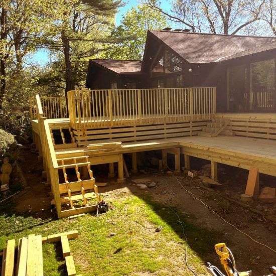 Wooden deck under construction beside a cabin in a wooded yard, with stairs and railings installed.