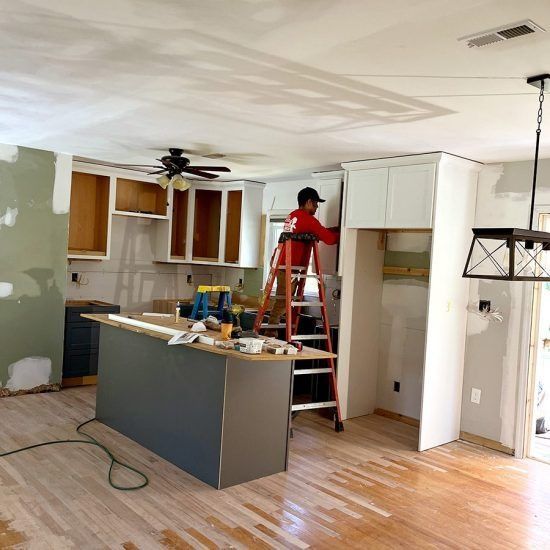 Worker on red ladder installing white kitchen cabinets in a partially renovated room.