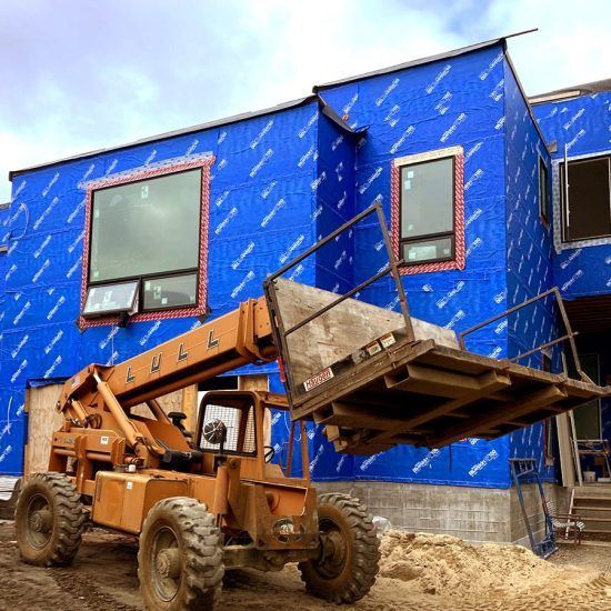 Orange skid steer lifting a wooden deck toward a blue house under construction
