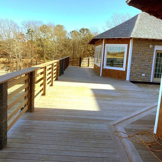 Wooden deck walkway beside a brick house, with railings and trees in the background.
