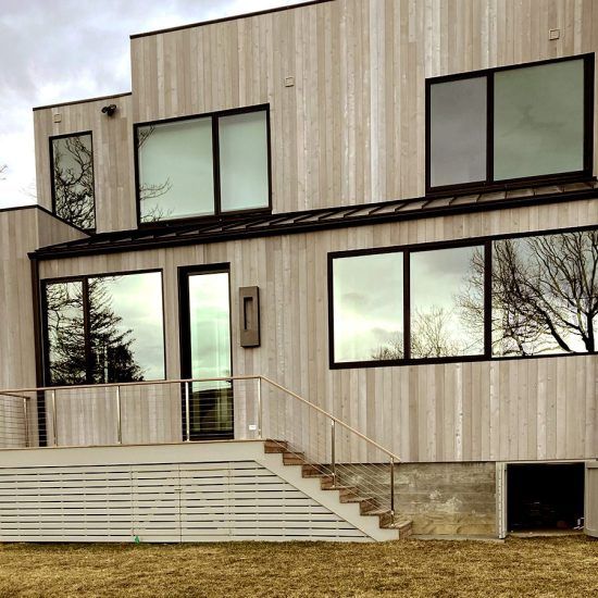 Modern two-story house with light wood siding, large windows, and a front deck with stairs and railing
