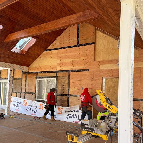 Workers installing siding on a house exterior under a covered porch, with a miter saw in the foreground
