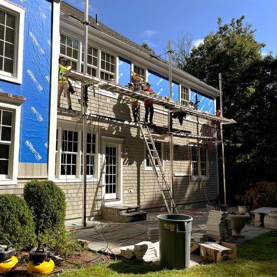 Workers on scaffolding repairing a two-story house exterior with blue siding and a ladder in the yard