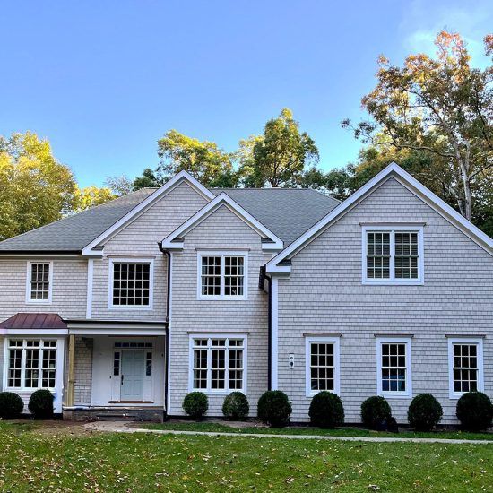 Large light gray suburban house with white trim, front porch, and manicured lawn under blue sky