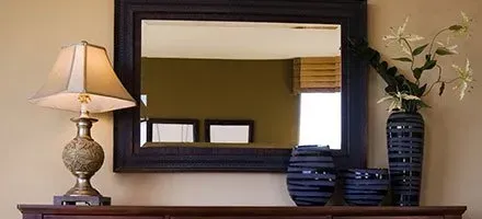 A dark-framed mirror on a wood surface, with a lamp and vase of flowers to the side.