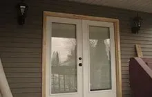 Double glass doors framed with wood on a brown siding exterior; two black sconce lights flank the doors.