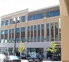 Three-story tan building with large windows and a storefront, street view with cars and trees.