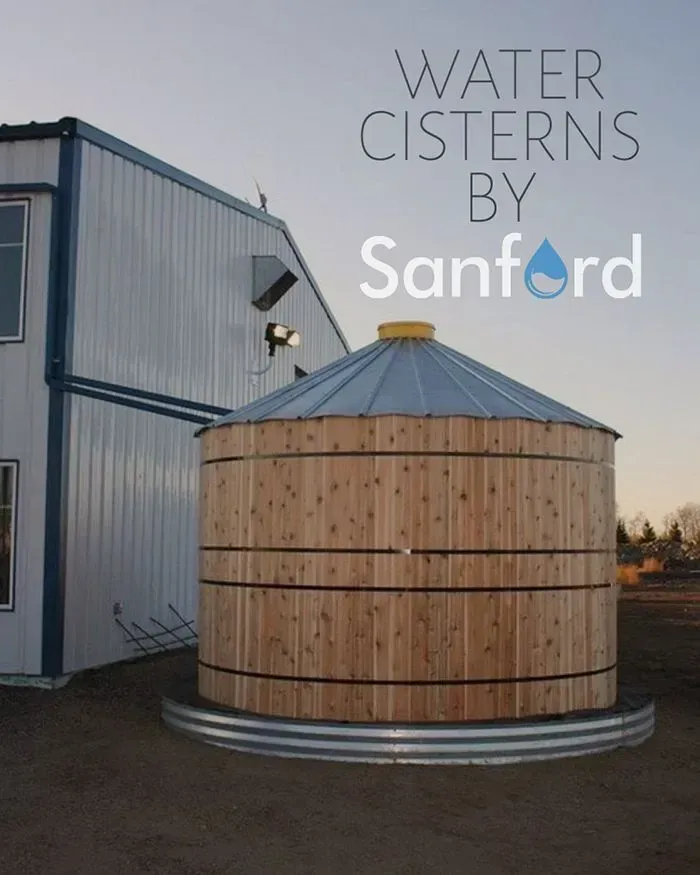Wooden water cistern by Sanford, next to a white building. The cistern has a metal roof and base.
