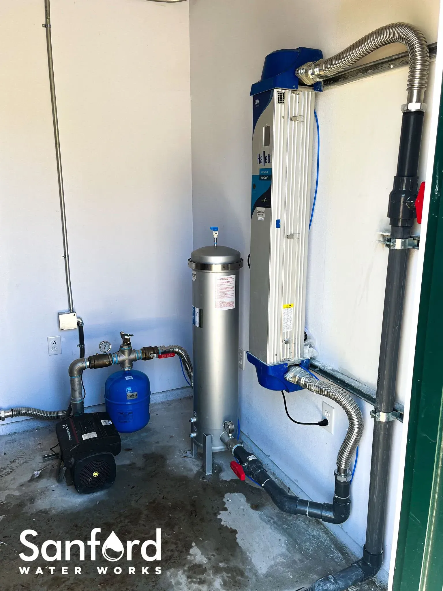 Water filtration system installed in a white utility room. The system has a blue pressure tank, silver cylinders, and black pipes.
