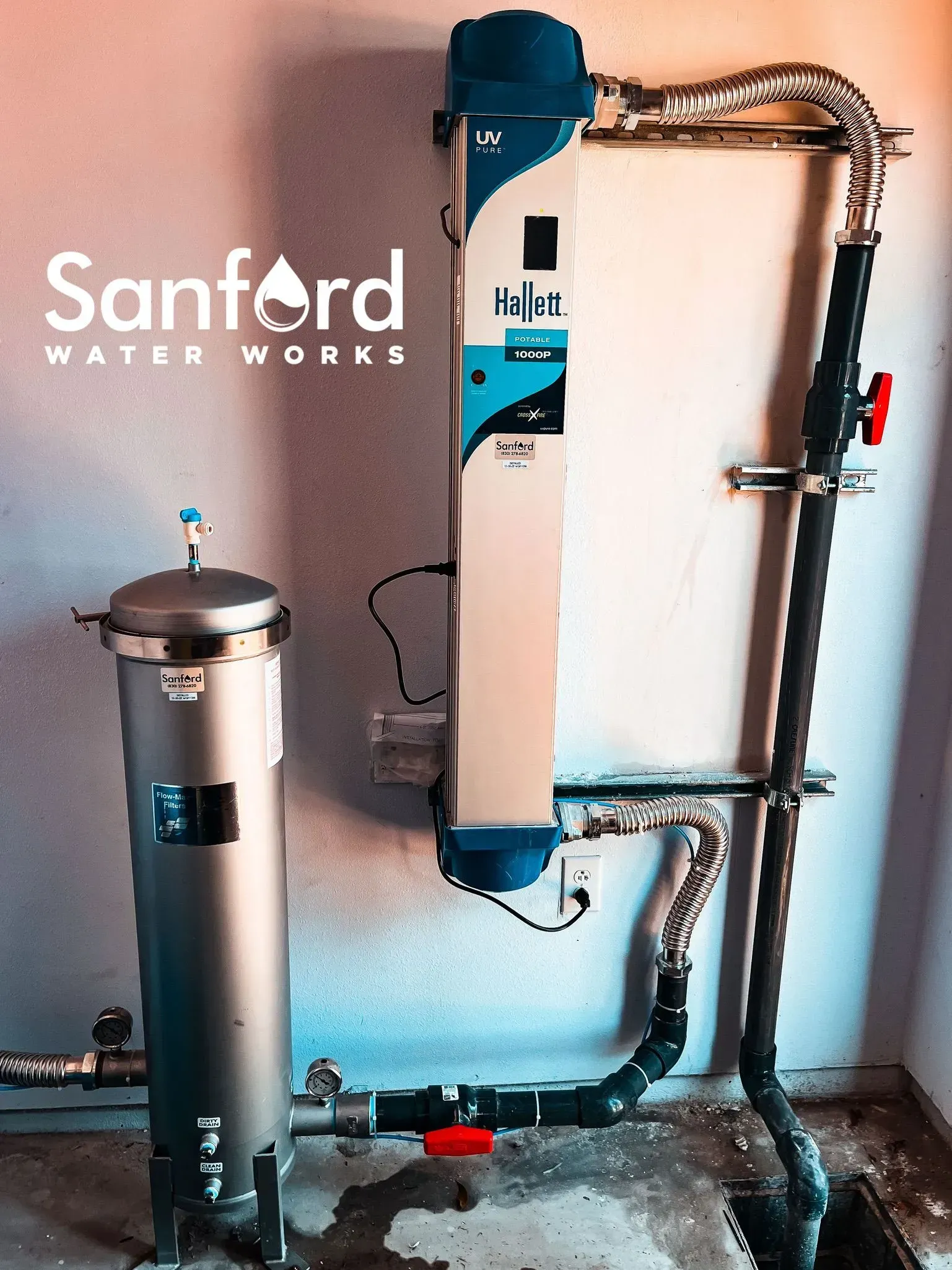 Sanford Water Works water filtration system, with two silver cylinders and connected pipes, installed against a wall.