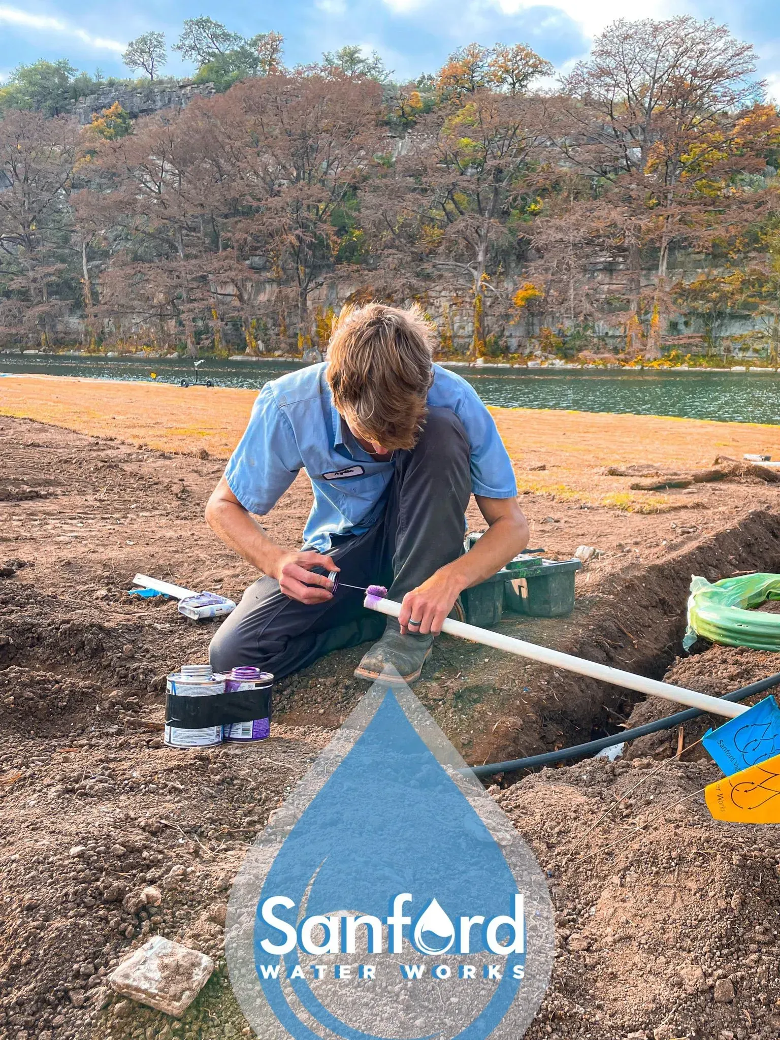 Man kneeling, connecting pipes outdoors near a water source, Sanford Water Works logo.