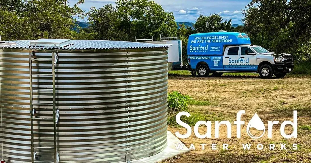 Galvanized water tank with a Sanford Water Works truck in a grassy field.