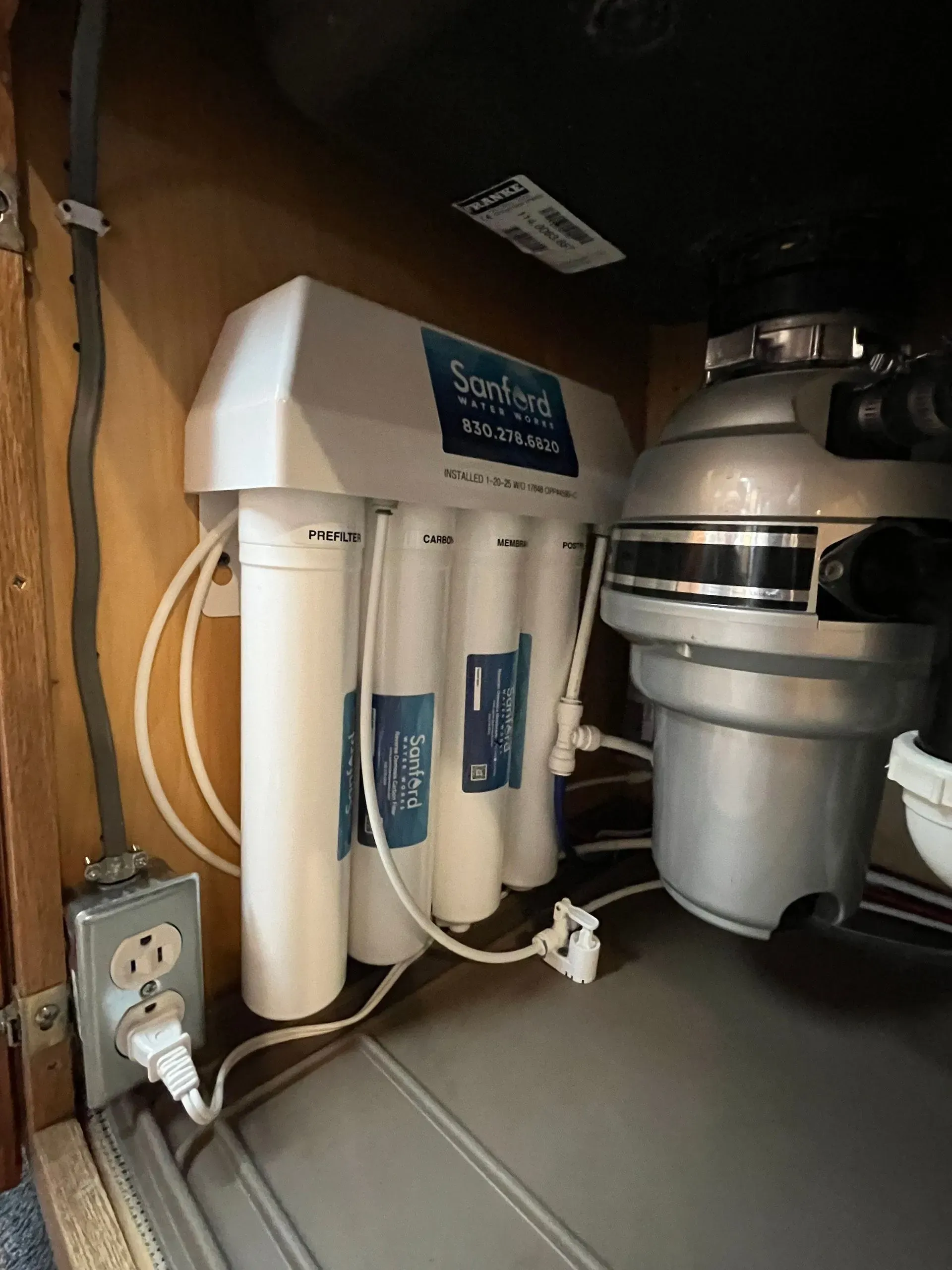 Under-sink water filter and garbage disposal unit, both with white and silver components, next to an electrical outlet.