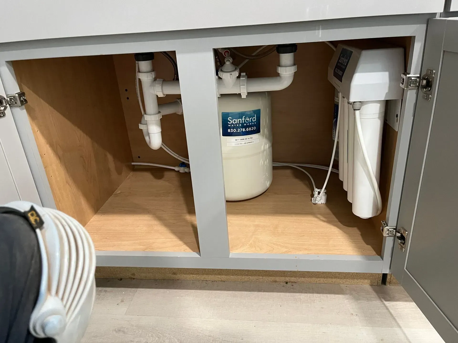 Cabinet under a sink with plumbing and a water filtration system. Grey and white colors are present.