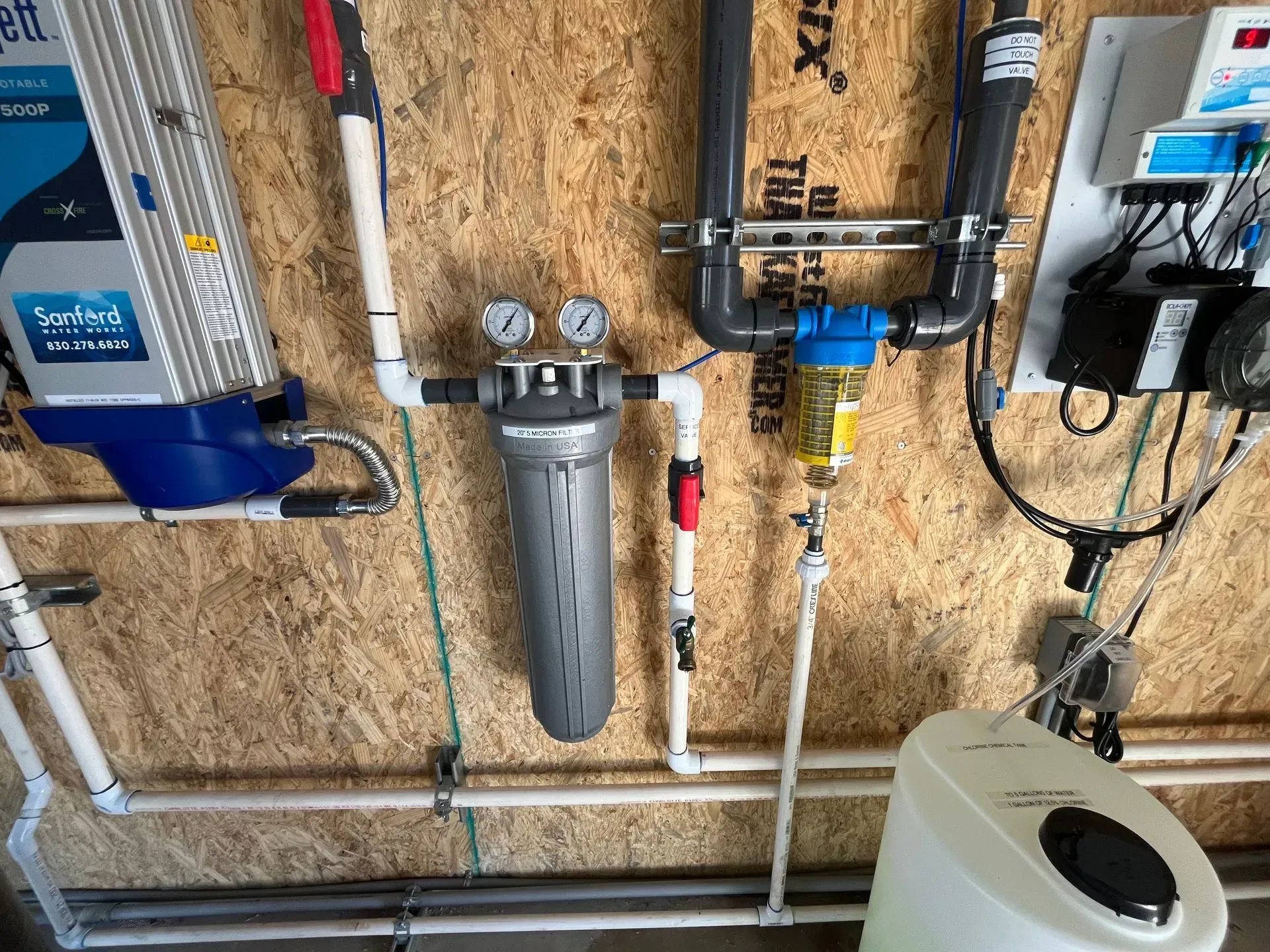 Water filtration system on a wooden wall, with pipes, gauges, and a softener unit.