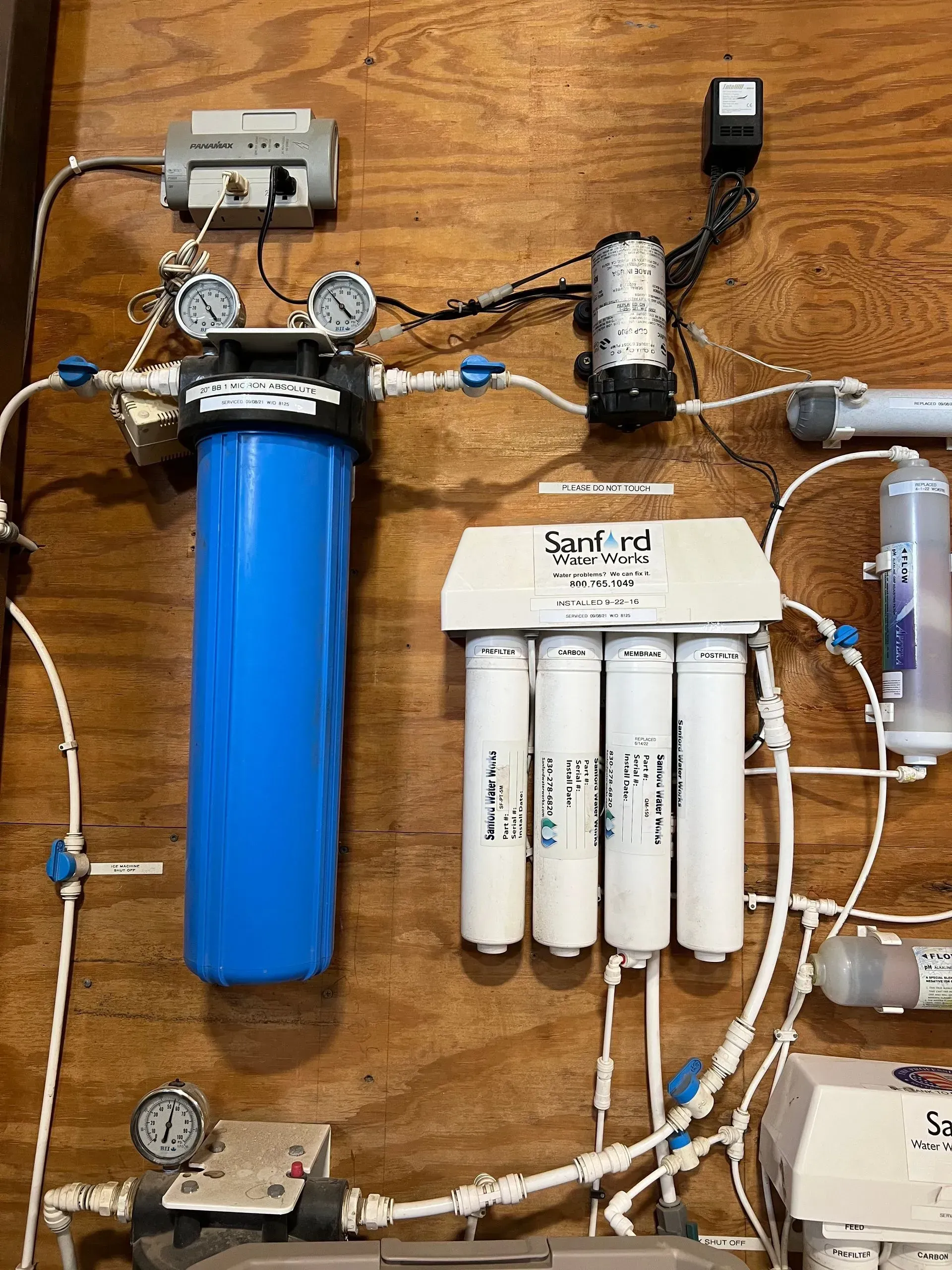 Water filtration system components mounted on a wood panel: blue filter housing, multiple white filters, gauges, pump.