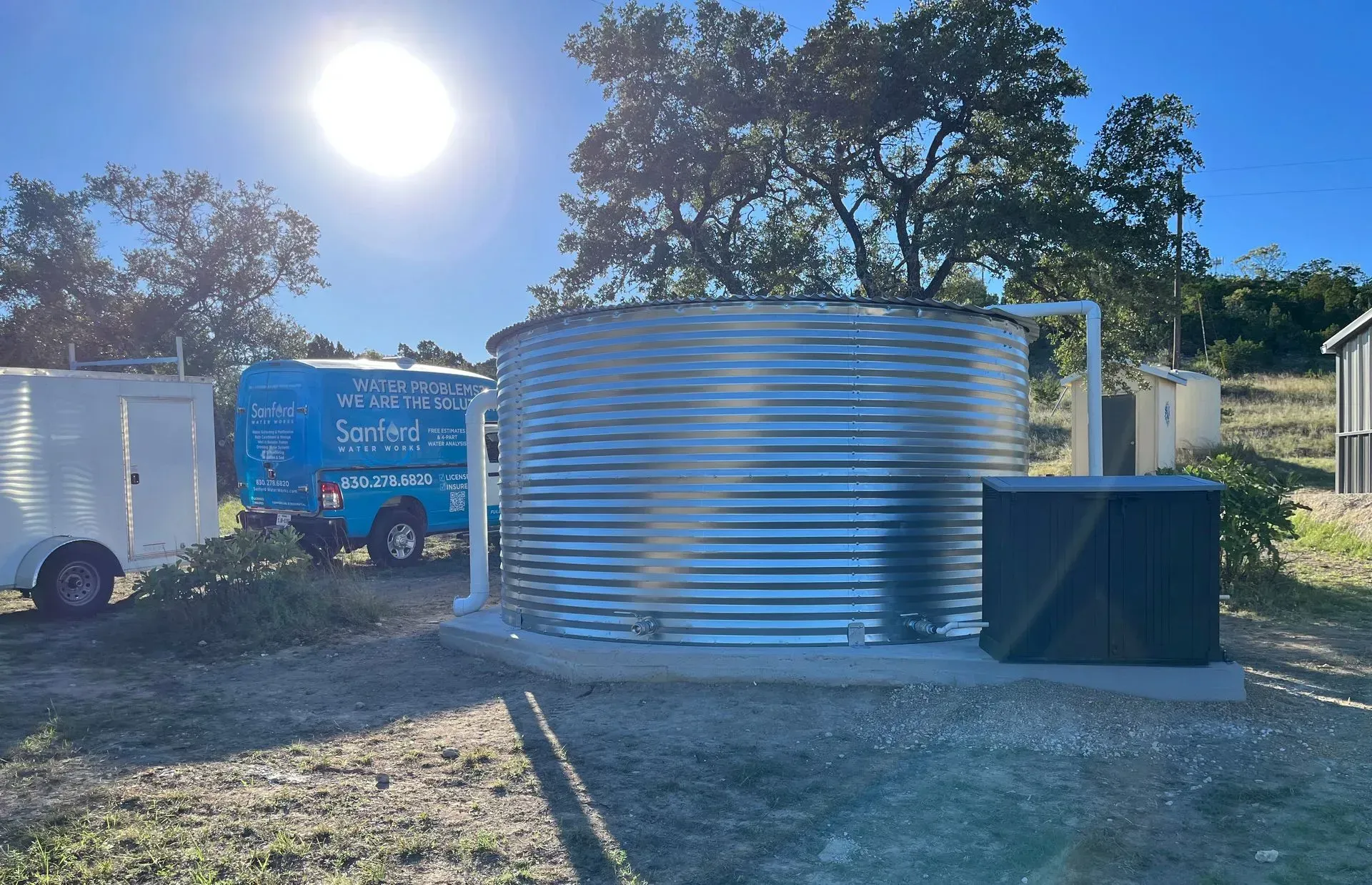 Large, corrugated metal water tank outside on a sunny day. Blue truck and small building are nearby.