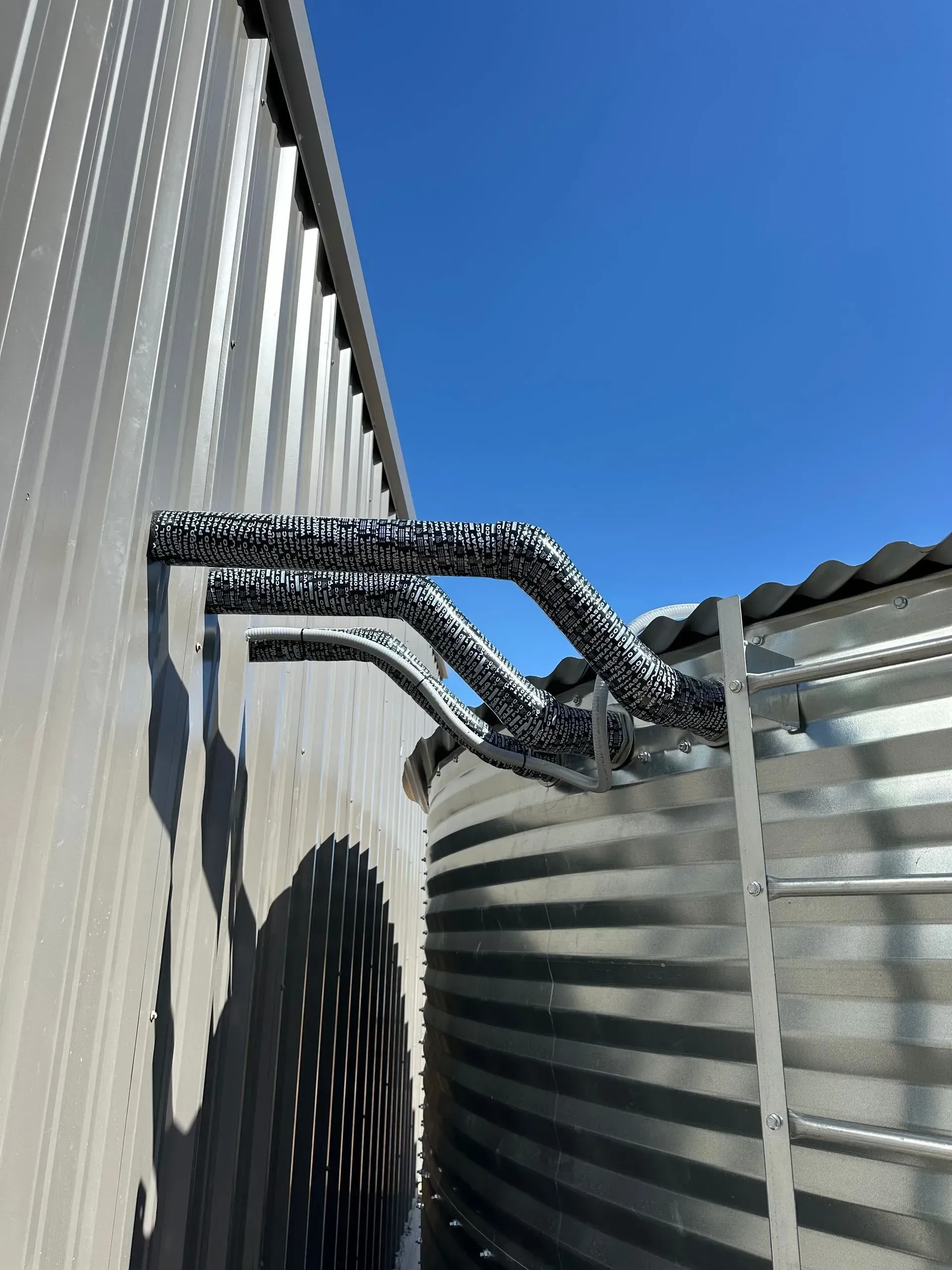 Pipes with insulation attached to the side of a metal building under a blue sky.