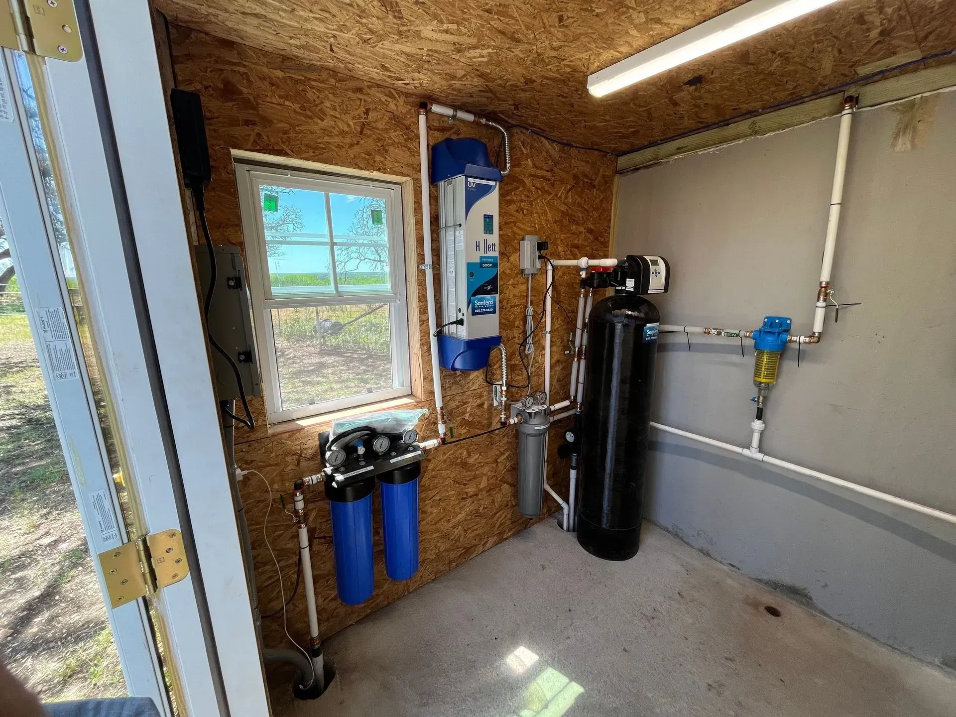 Water filtration system in a small shed with multiple tanks and filters, connected by white pipes.