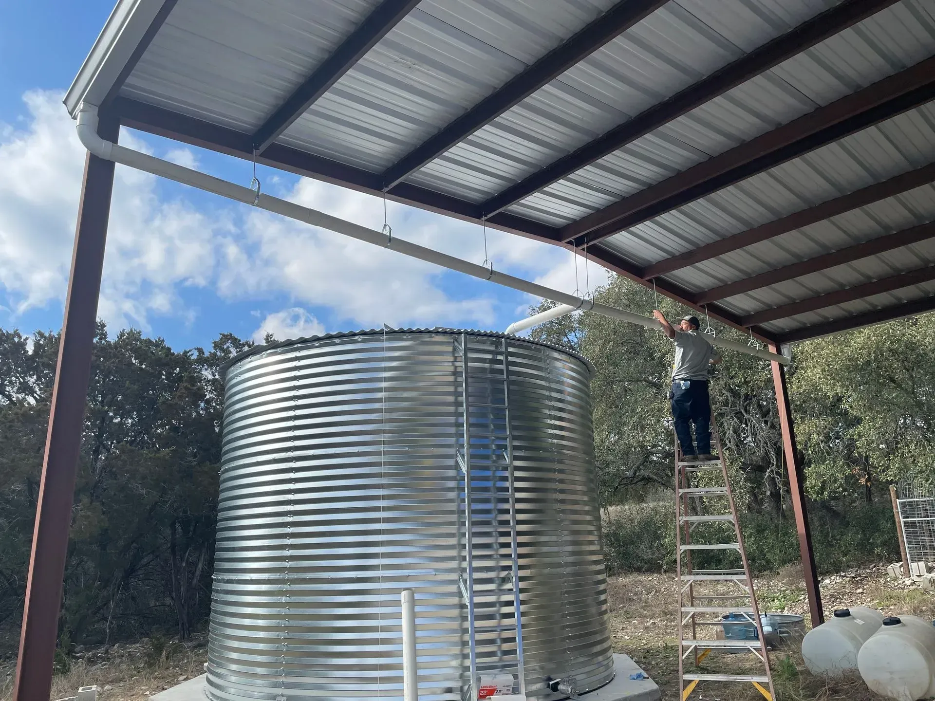 Person on ladder installing gutter over large metal water tank. Shelter overhead.