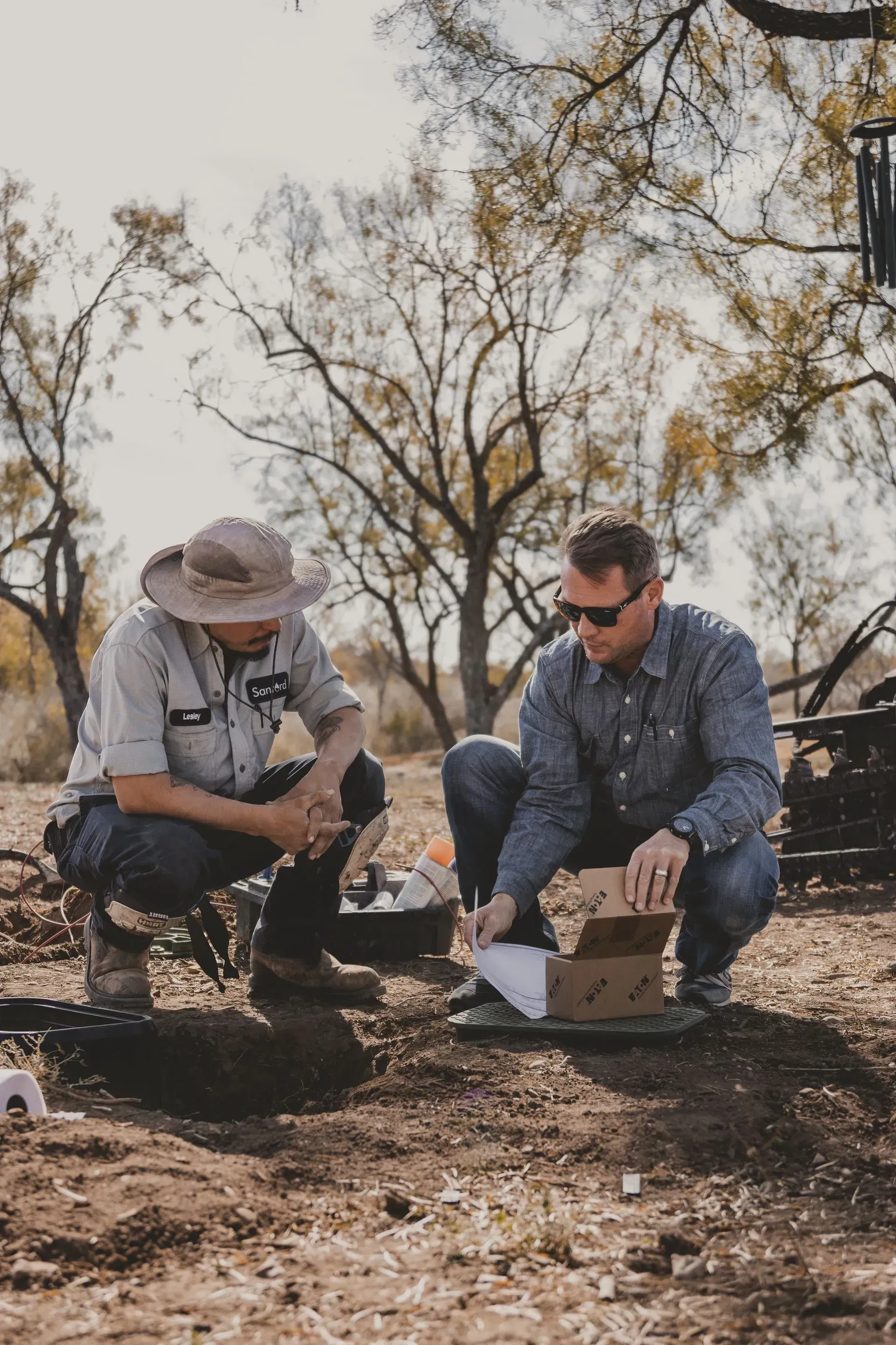 Two men squat outside, examining papers from a box. Dry, brown ground. Trees in the background.